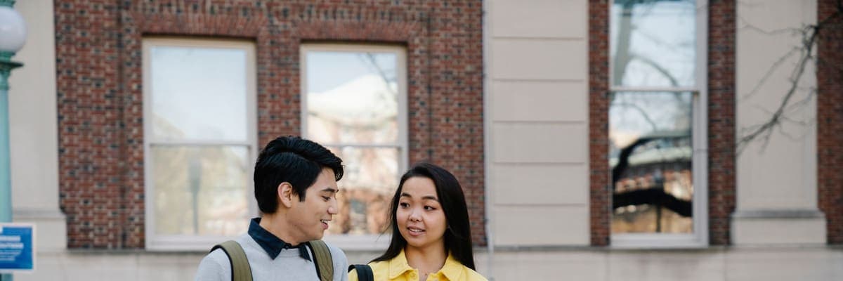 Two students walking outdoors past a campus building, carrying bags.